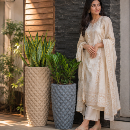 A woman in a cream outfit stands beside two tall diamond-textured planters holding green plants.